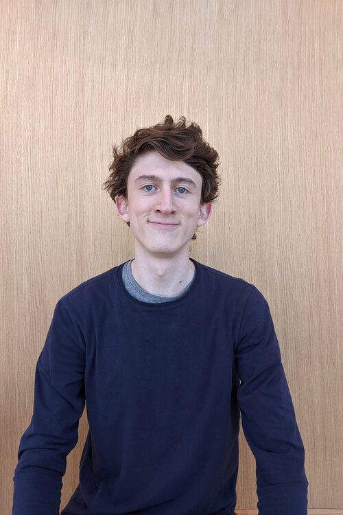 Photo of Corbin Auriti Owner of No BS Technologies in front of a wooden background wearing a blue shirt